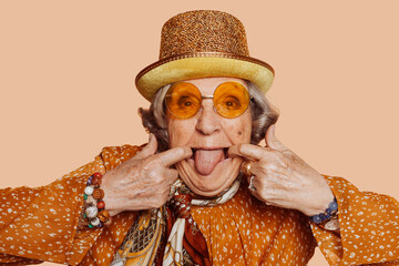 Close up portrait of elderly stylish grandmother doing funny face at studio over beige background. Comedian face, with fingers opening mouth showing tongue.