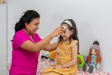 Latin female pediatrician checking the temperature of a girl with a dress, sitting on the stretcher in the office