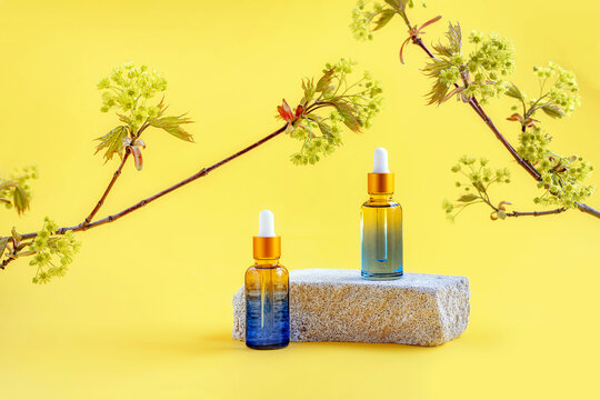 bottles with a pipette on a podium made of stone and branches of a flowering maple on a yellow background, selective focus