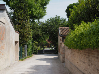 Gravel road between brick buildings leading to a garden house. Closed gate.