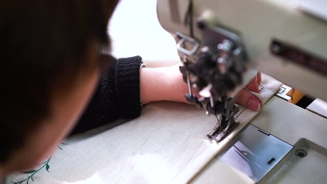 The woman begins to stitch on the fabric with overhanging floral ornaments. With his hand, he straightens and stretches out the fabric. An industrial sewing machine stitches a stitch