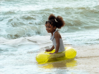 Cute little African girl wearing striped swimsuit with yellow inflatable swim ring playing with wave. The girl having fun and happiness playing with splashing wave on the beach.