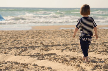 Little boy on the beach looks at the sea. A child in a sailor's T-shirt.  Back view. Concept: travel dreams, summer family vacation