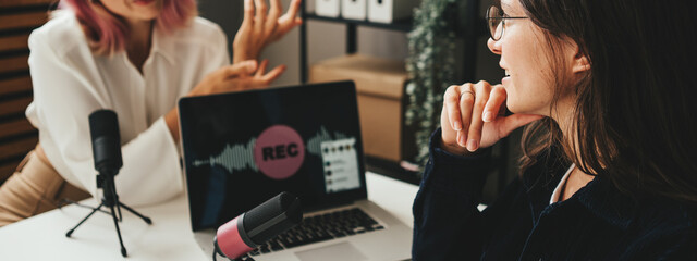 Two friendly women host streaming audio podcast using microphone and laptop at home broadcast studio