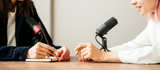 Two woman hosts in local broadcat studio recording audio podcast, hands close-up, sitting opposite...