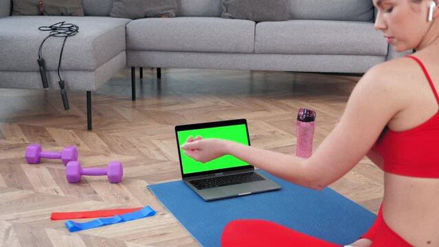 Fit woman in sportswear sits on yoga mat looks computer display, tells fitness trainer online video call webcam, distance sports lesson course. Green screen mock up chroma key monitor laptop concept