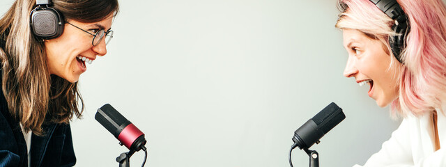 Two women in local broadcat studio recording audio podcast. Sitting opposite each other, smiling