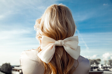 Back view of woman with long fair hair decorated with beige bow made of ribbon standing on blue sky background. Close-up