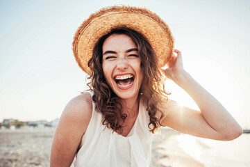 Young joyful woman in white shirt wearing hat smiling at camera on the beach - Traveler girl...