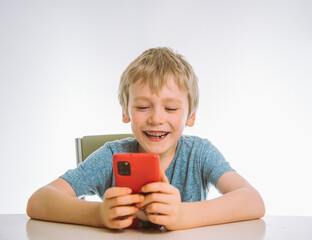 the boy holds a smartphone in his hands and looks at his screen emotionally with a smile. sitting at a table, white background.