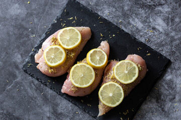 Raw chicken breast fillets, covered with rosemary and sliced lemon, on a slate cutting board.  On a dark stone background