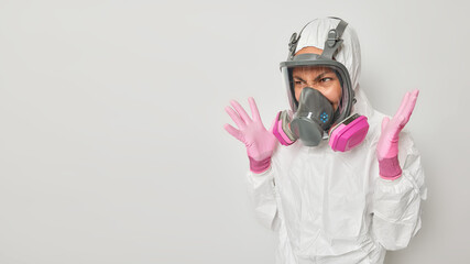 Outraged woman dressed in protective suit with hood wears gas mask keeps palms raised realizes being under threat in difficult situation isolated over white background blank space for promo.