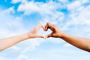 Two multiethnic hands join together to form a shape heart with a blue sky clouds background....