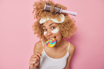 Lovely curly haired young woman bites multicolored lollipop has sweet tooth applies beauty patches...