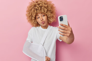 Injured woman with curly hair suffers from pain wears sling on arm keeps lips folded takes selfie via smartphone keeps lips folded dressed in casual white t shirt isolated over pink background