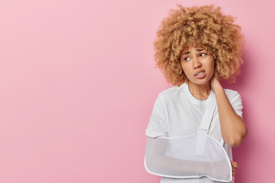 Unhappy curly haired young woman has broken arm wears sling frowns face looks away keeps hand on neck dressed in casual white t shirt isolated over pink background blank space for your promotion