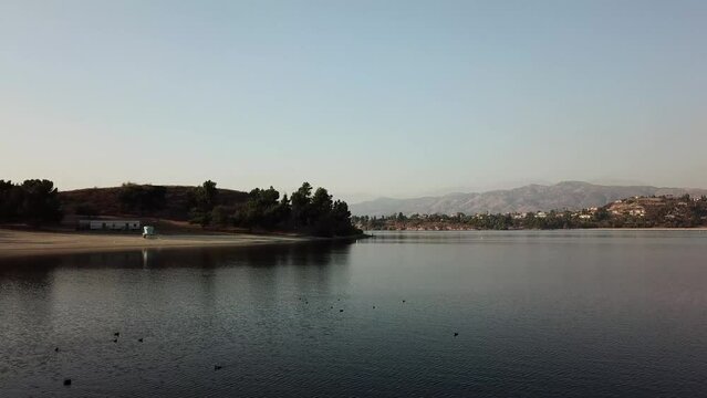 aerial footage of a vast blue rippling lake at sunset surrounded by lush green trees, grass and plants with majestic mountain ranges and blue sky at Puddingstone Lake in San Dimas California USA