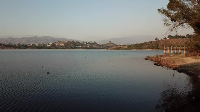 aerial footage of a vast blue rippling lake at sunset surrounded by lush green trees, grass and plants with majestic mountain ranges and blue sky at Puddingstone Lake in San Dimas California USA