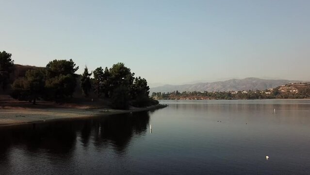aerial footage of a vast blue rippling lake at sunset surrounded by lush green trees, grass and plants with majestic mountain ranges and blue sky at Puddingstone Lake in San Dimas California USA