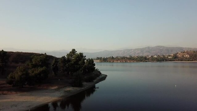 aerial footage of a vast blue rippling lake at sunset surrounded by lush green trees, grass and plants with majestic mountain ranges and blue sky at Puddingstone Lake in San Dimas California USA