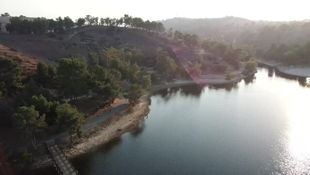 aerial footage of a vast blue rippling lake at sunset surrounded by lush green trees, grass and plants with majestic mountain ranges and blue sky at Puddingstone Lake in San Dimas California USA