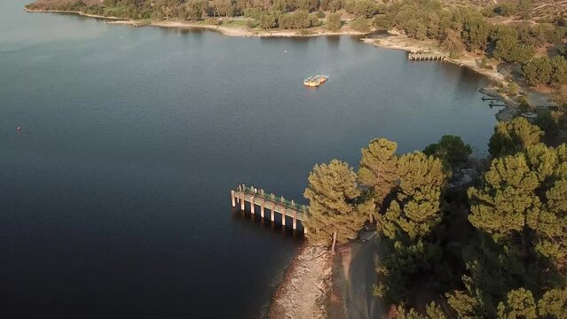 aerial footage of a vast blue rippling lake at sunset surrounded by lush green trees, grass and plants with majestic mountain ranges and blue sky at Puddingstone Lake in San Dimas California USA