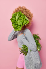Unrecognizable young woman with curly hair covers face with green fresh salad carries bag full of fresh vegetables dressed in sportswear isolated over pink background. Healthy eating concept
