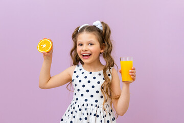 A little girl holds a glass of orange juice and a slice of orange, freshly squeezed juice for...