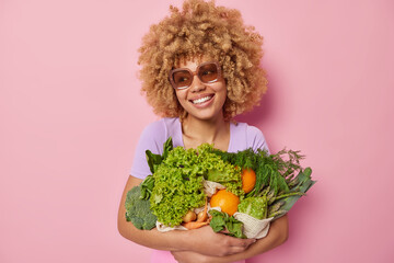 Cheerful woman wears sunglasses embraces bunch of fresh green vegetables picks up grocery from own...