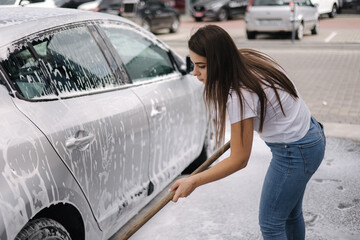 Attractive young woman washing her car with shampoo and brushes. Female washes automobile with foam and water outside on self service car wash