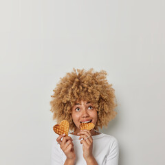 Vertical shot of lovely curly haired woman bites appetizing heart shaped waffles focused above...