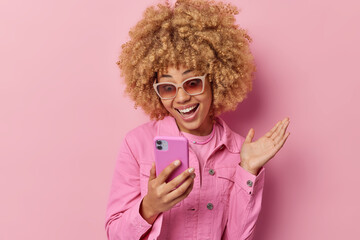Positive fashionable young woman with curly hair focused at smartphone gets excellent news in...