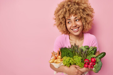 Cheerful curly haired woman embraces green fresh vegetables grown in own garden eats healthy...