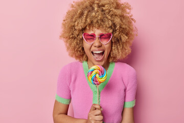 Overjoyed curly haired young woman laughs happily keeps mouth opened holds rainbow lollipop on...