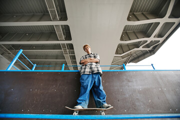 Low angle portrait of teenage boy wearing baggy pants standing on skateboard in urban area and...