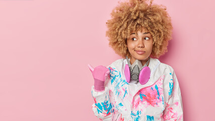 Curious young woman with curly hair wears safety suit and rubber gloves points thumb away on copy space tells you about biochemical hazard and virus protection isolated over pink background.