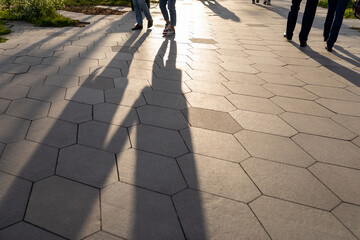 The feet of strolling pedestrians on the sidewalk paved with hexagonal large tiles. Long shadows on...