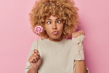 Funny curly haired woman makes grimace keeps eyes crossed shows arm with elastic bandge after getting into accident holds caramel lollipop dressed in casual t shirt isolated over pink background
