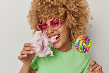 Cheerful curly haired woman bites delicious candy floss with appetite holds multicolored candy on stick enjoys eating appetizing treats wears heart shaped sunglasses green t shirt. Summer lifestyle