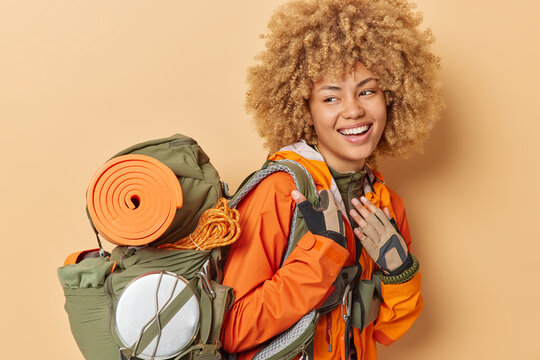 Positive woman with curly bushy hair has camping travel trip dressed in orange windbreaker carries rucksack leads active lifestyle isolated over beige background. Female traveler prepares for hking