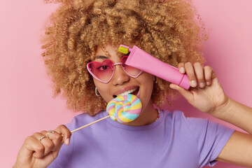 Horizontal shot of curly haired European woman bites lollipop holds toothbrush and toothpaste wears trendy sunglasses casual purple t shirt isolated over pink background. Harmful food concept