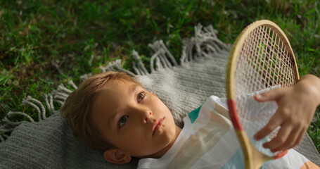 Closeup upset boy examining wooden badminton racket lying green grass alone