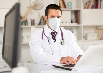 Focused male doctor in medical face mask listening to patient complaints and writing prescription at clinic