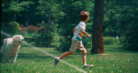 Active little boy running playing with dog on field with sprinklers on sunny day - obrazy, fototapety, plakaty