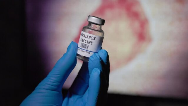 doctor holding a vial of smallpox vaccine in front of the monkeypox virus graphic on a monitor