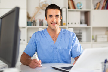 Portrait of smiling experienced physician filling up medical forms on laptop while sitting at table in office