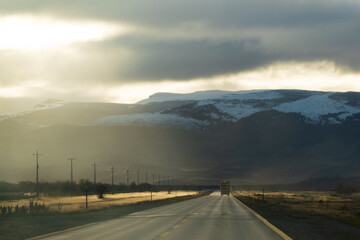 carretera asfaltada alumbrada por rayos de sol al atardecer saliendo de entre las nubes nimbostrato con cerros altos  nevados de fondo y un camión de carga a lo lejos 