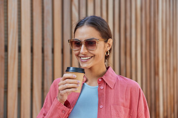 Beautiful cheerful woman with dark hair drinks coffee from disposable cup enjoys fresh hot beverage wears trendy sunglasses casual pink shirt smiles positively poses against wooden hence background