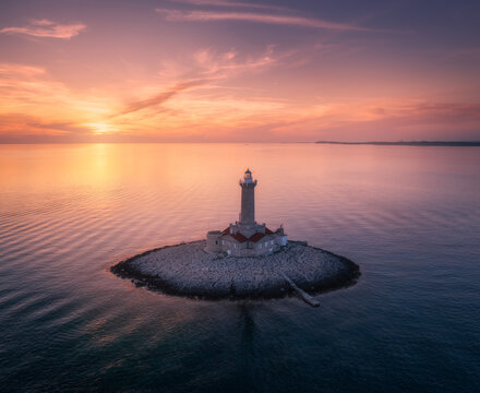 Lighthouse on smal island in the sea at colorful sunset in summer. Aerial top view of beautiful lighthouse on the rock, water and orange sky with pink clouds at dusk. Landscape. Adriatic sea, Croatia