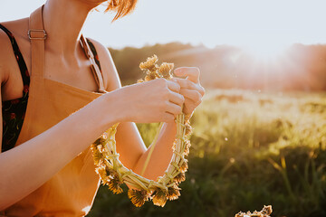 Portrait of beautiful woman making wreath of flowers dandelions on flowering field. Summer...